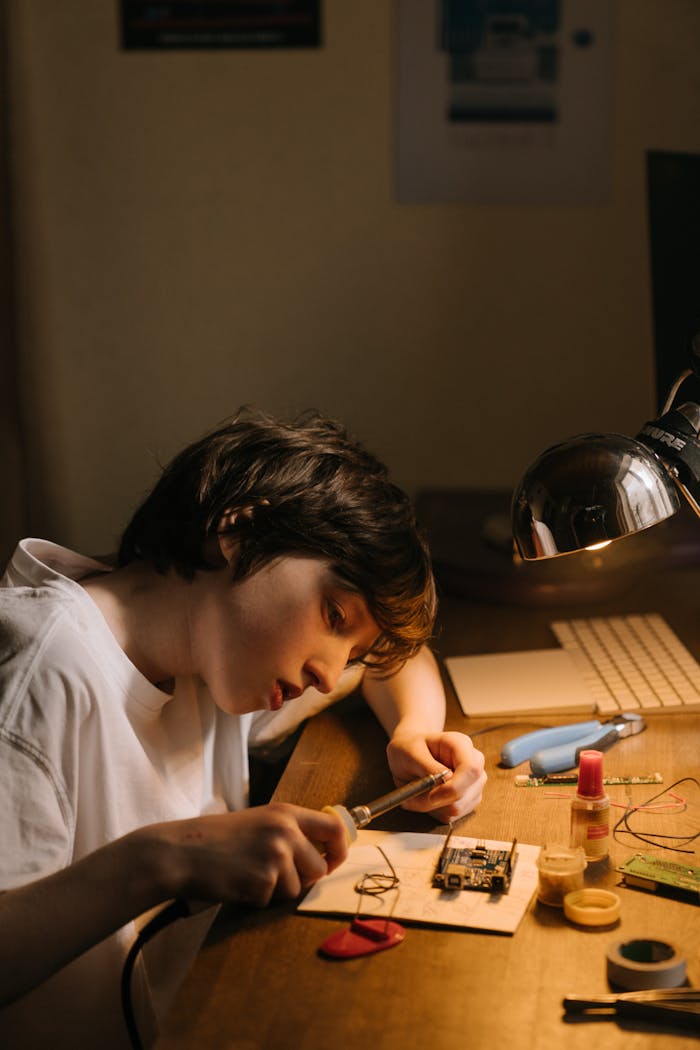 services-01 A boy focused intently on repairing a circuit board using a soldering iron at his desk.